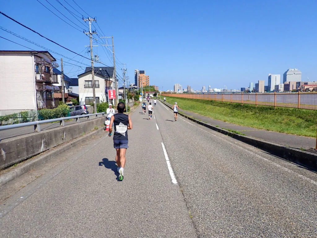 遠くに高層ビルが見える川沿いの道を、ゴールへ向かって走るランナー