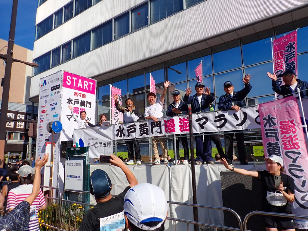 「START」と「水戸黄門漫遊マラソン」の看板を掲げたスタート台で手を振る人たち