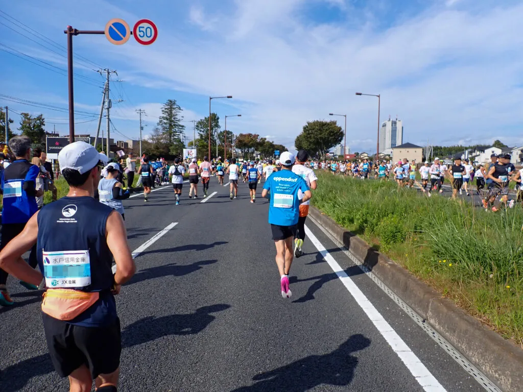 広い道路いっぱいに広がって走る参加者と、土手の上の沿道