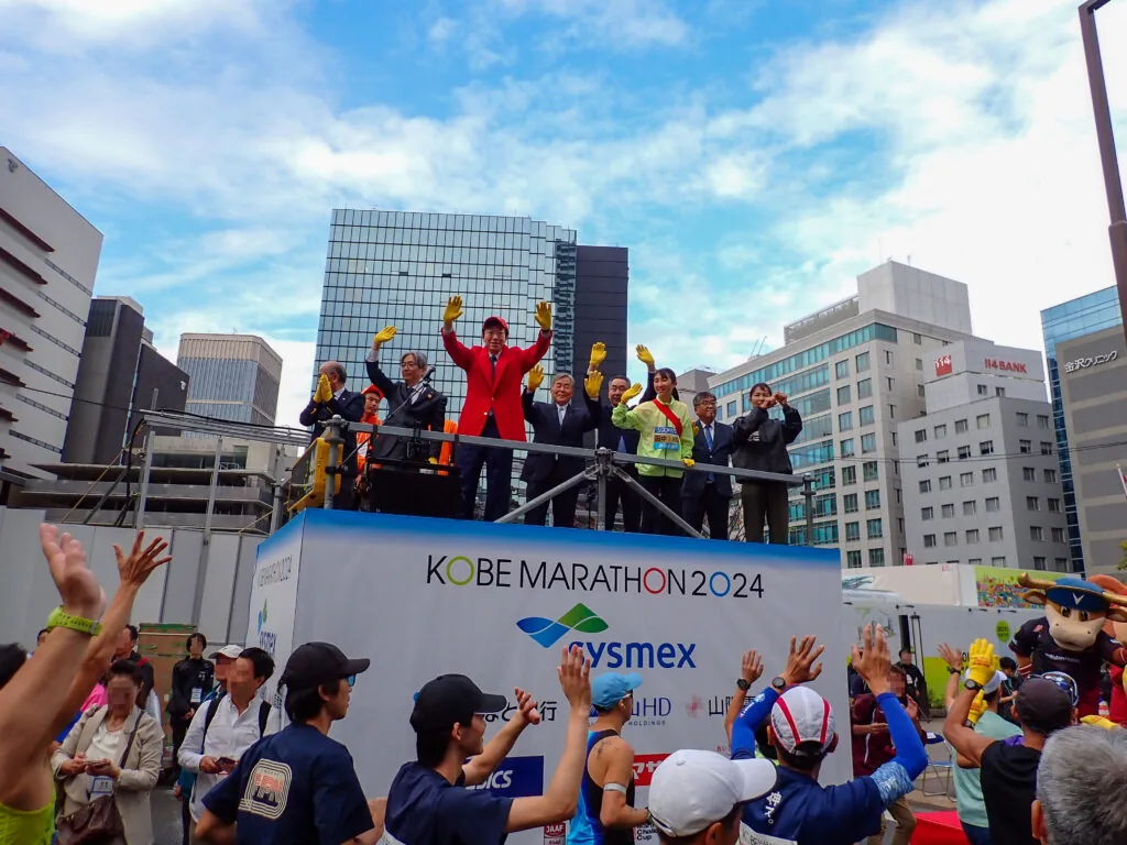「KOBE MARATHON」の看板を掲げたスタート台で手を振る人たち