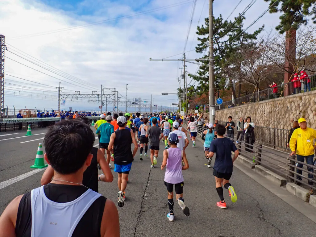 線路の架線が見える海沿いの道を走るランナーたちと、右の石垣の上で応援する人たち