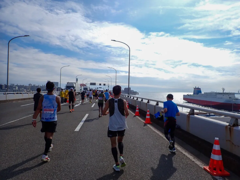 海に浮かぶ大きな船を横に見ながら、橋の上を走るランナーたち