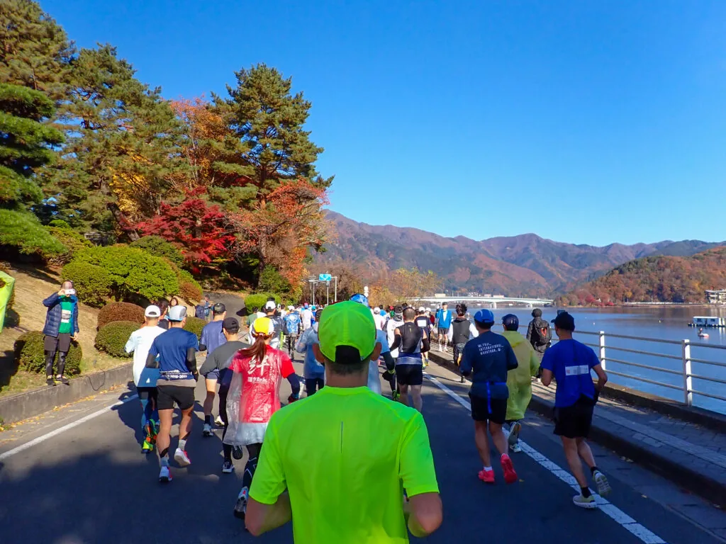 紅葉の木々と山並みを望む湖畔の道を、河口湖大橋の方へ走る参加者たち