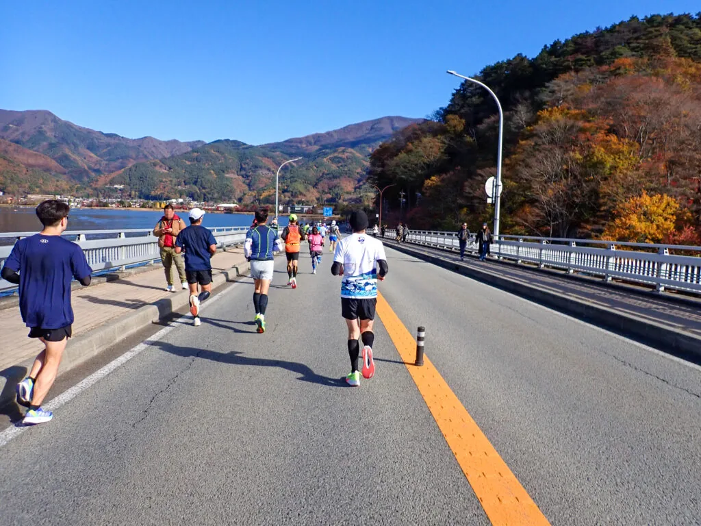 湖と山並みを望む河口湖大橋を渡り、紅葉の斜面を右手に走るランナーたち