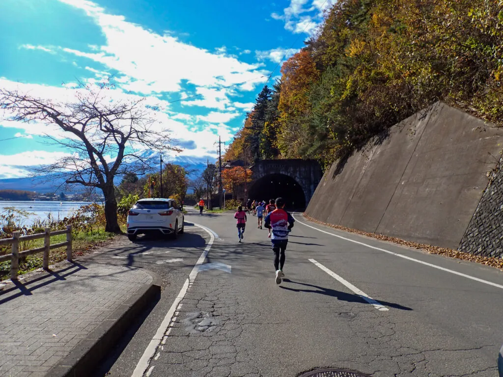 湖沿いの道から、紅葉の斜面に口を開ける長崎トンネルへ向かうランナーたち