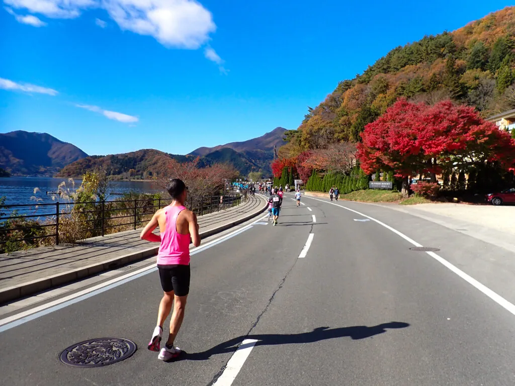真っ赤な紅葉と湖、山並みを見渡せる湖畔の道を走るランナー