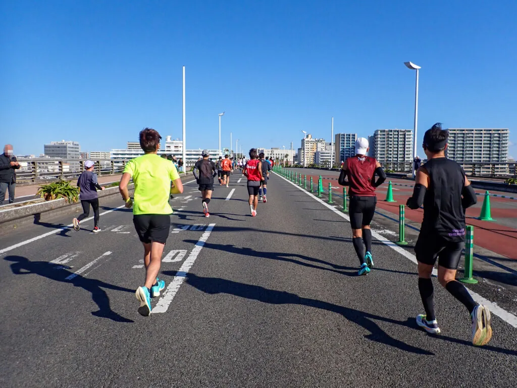 遠くに集合住宅が並ぶ、大淀川にかかる橋の上を走るランナーたち