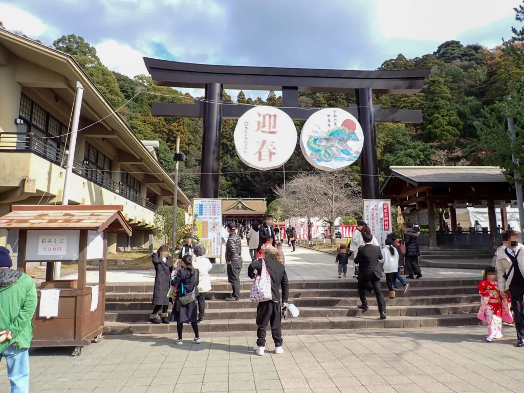 「迎春」の大きな丸い飾りが掛かる黒い鳥居と、石段を上る参拝の人たち