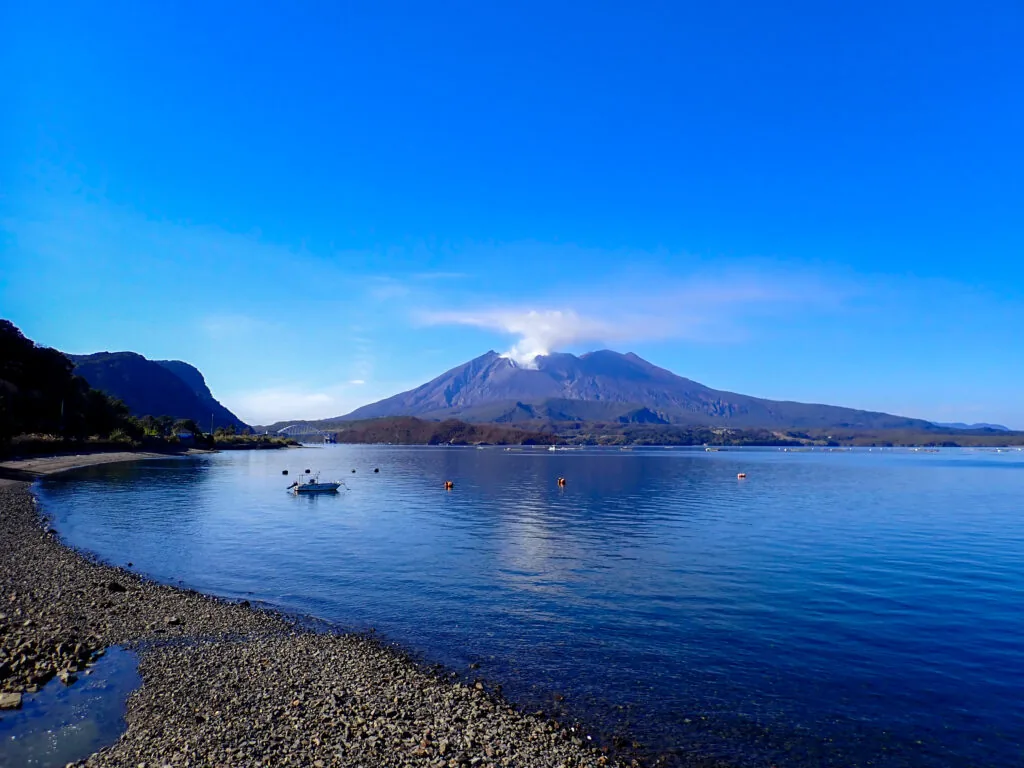 石の浜辺と穏やかな海の向こうに、噴煙を上げる桜島