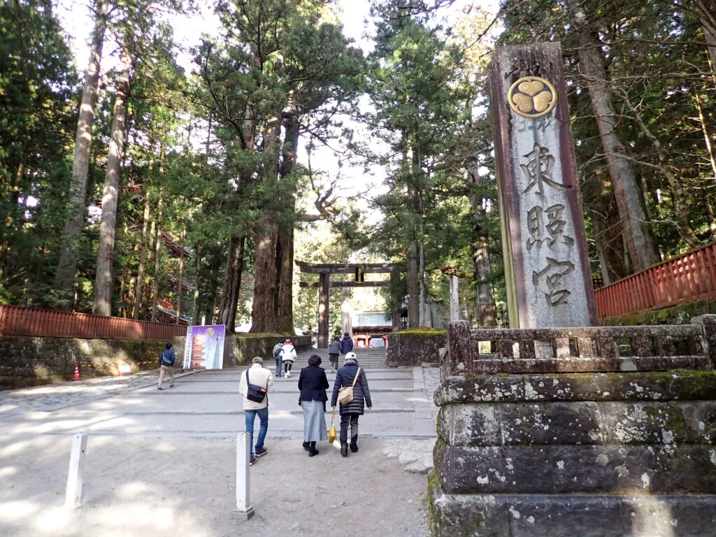 三つ葉葵の紋を載せた「東照宮」の社号標と、杉木立の参道を歩く人たち