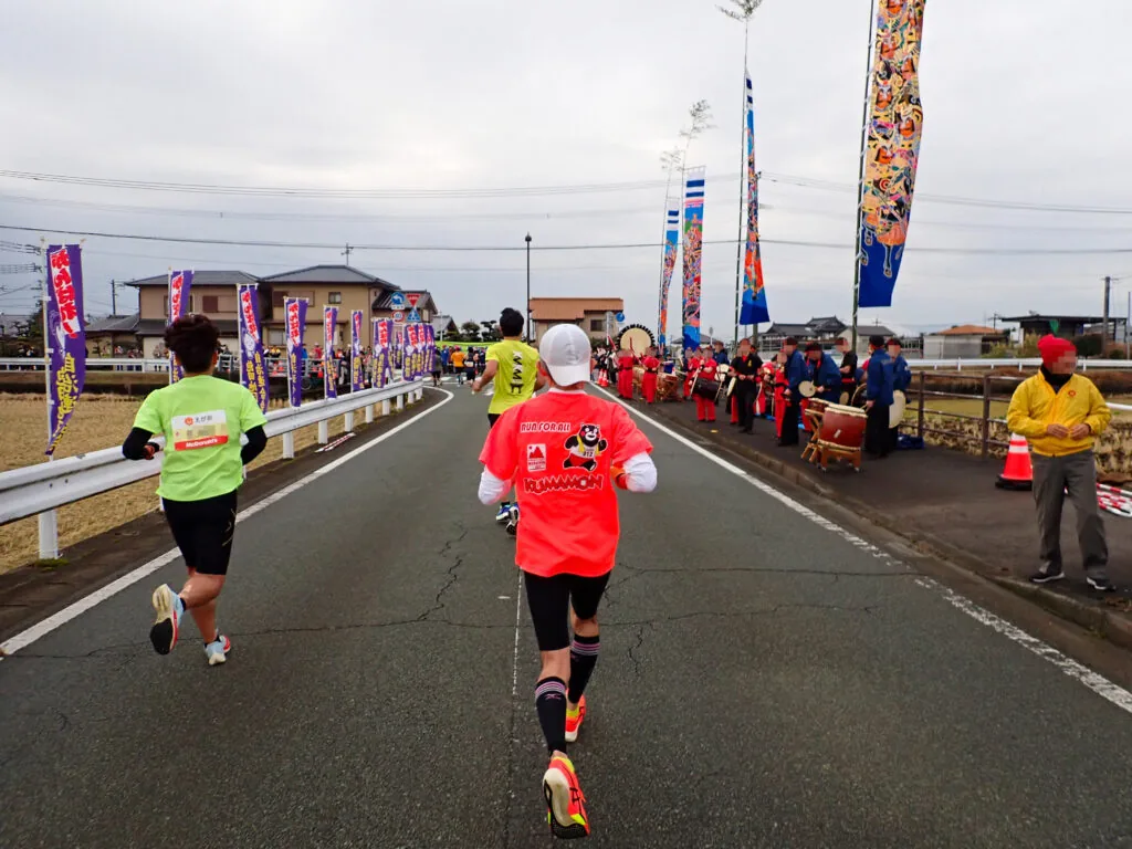 のぼり旗が並ぶ沿道で太鼓の応援を受けるランナーたち