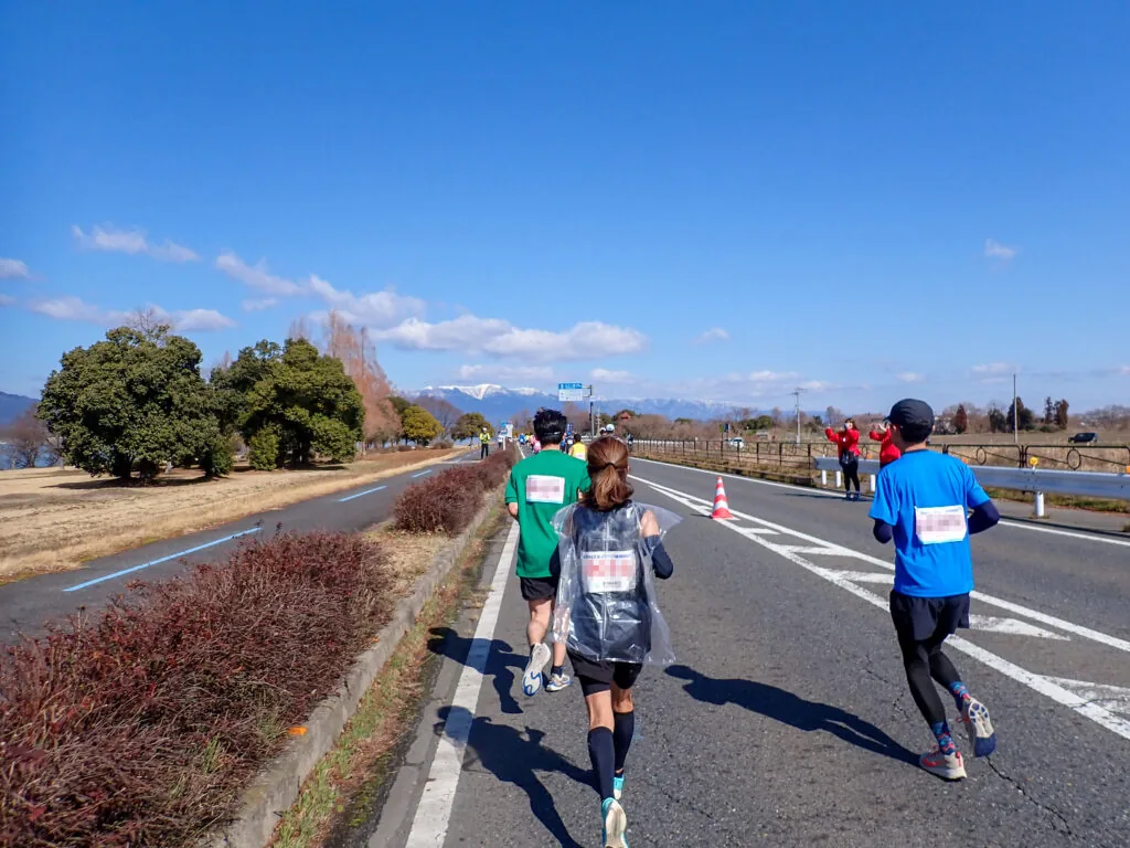 雪をかぶった山並みを望む湖岸道路を走るランナーたち