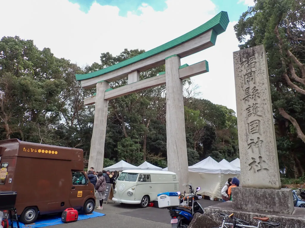 緑の笠木の鳥居と護国神社の石柱、手前に並ぶ出店の車